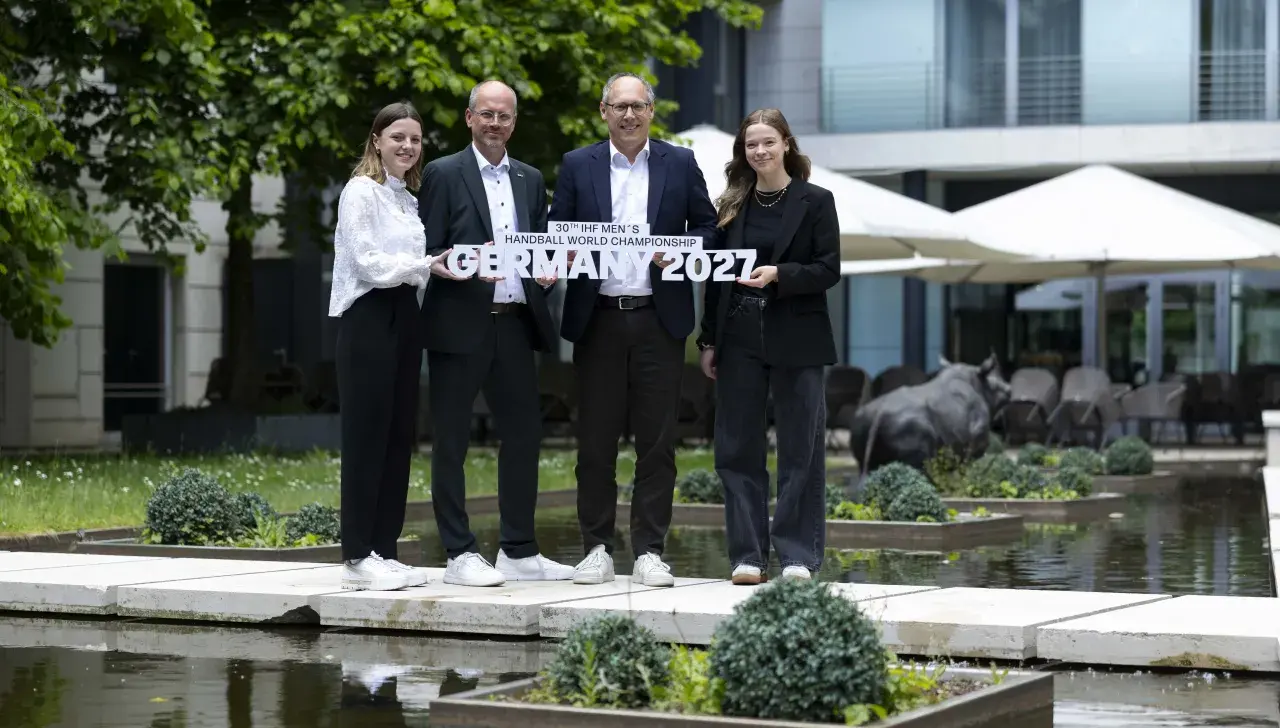 Vier Personen halten vor dem Radisson Hotel einen Schriftzug mit dem Namen der Handball-WM 2027 in den Händen.